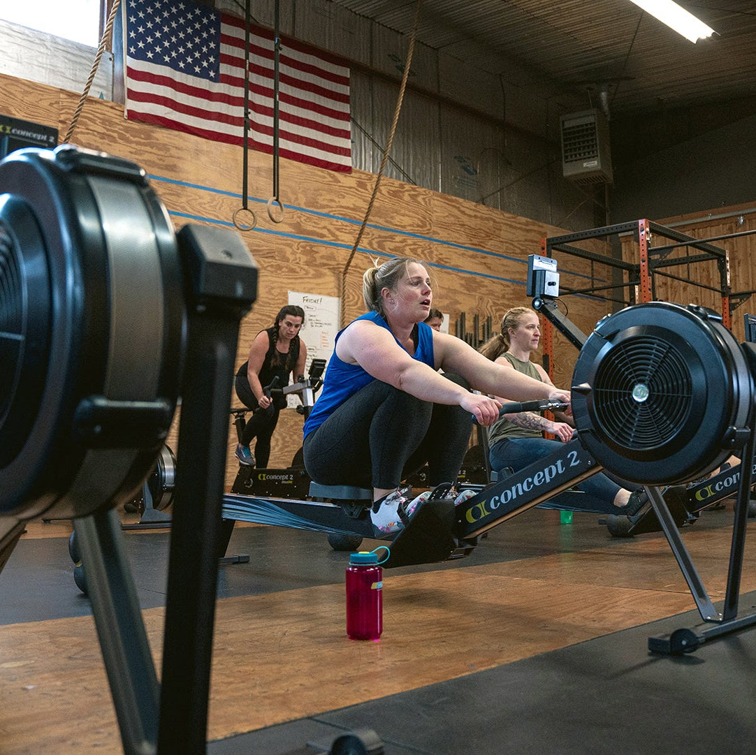 People using rowing machines in a gym with an American flag on the wall.