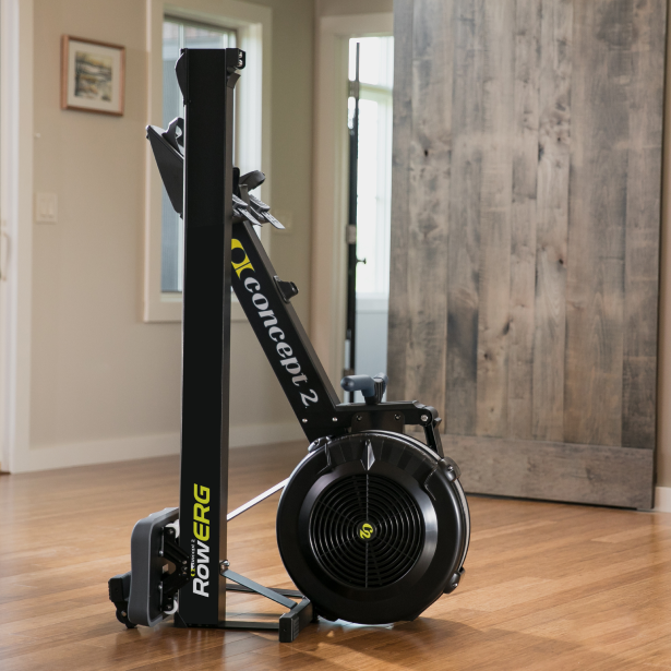 Concept II rowing machine in a home setting with wooden flooring and a wall-mounted mirror.